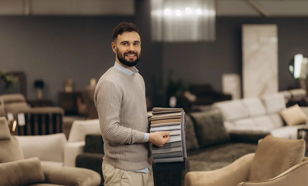 Mann im hellen Pullover hält Fächer mit Polsterstoff Mustern in Beige und Grau, im Hintergrund Sofas und Sessel in einer Möbel Ausstellung.